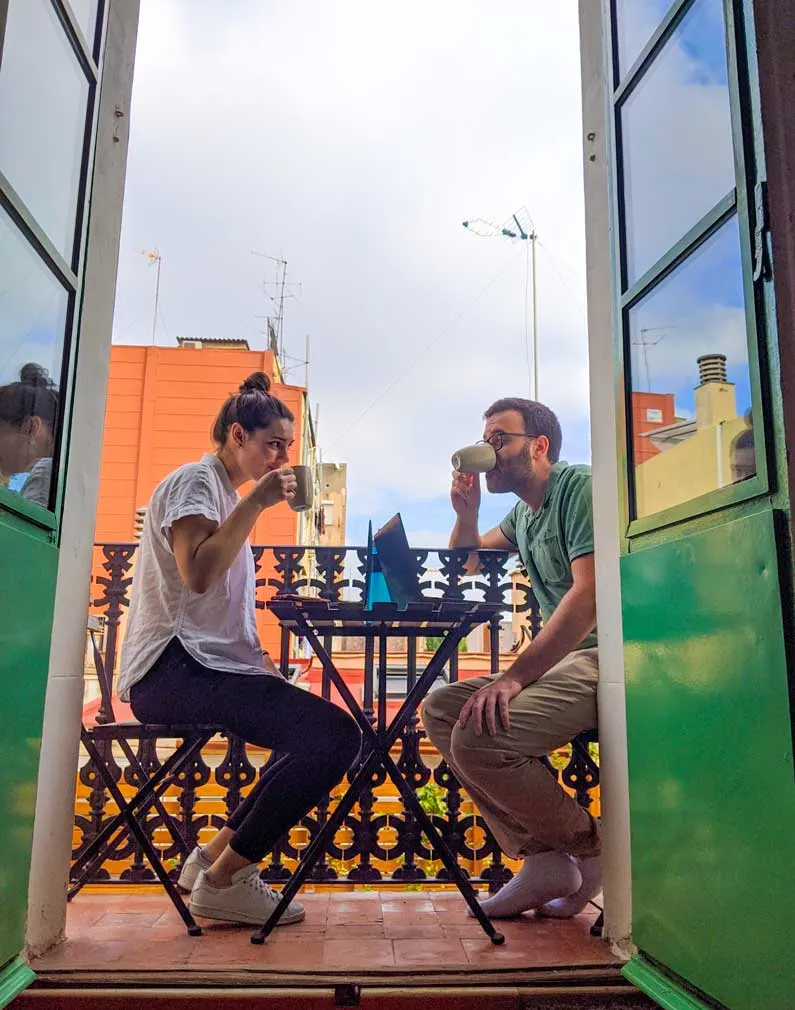 a vertical portrait giving viewers an insiders look at a couple remote work and travel life. Green doors open up onto a tiny balcony. A couple sits on a small ikea table, looking at each other, and each drinking their coffee, in front of each of them is their laptop