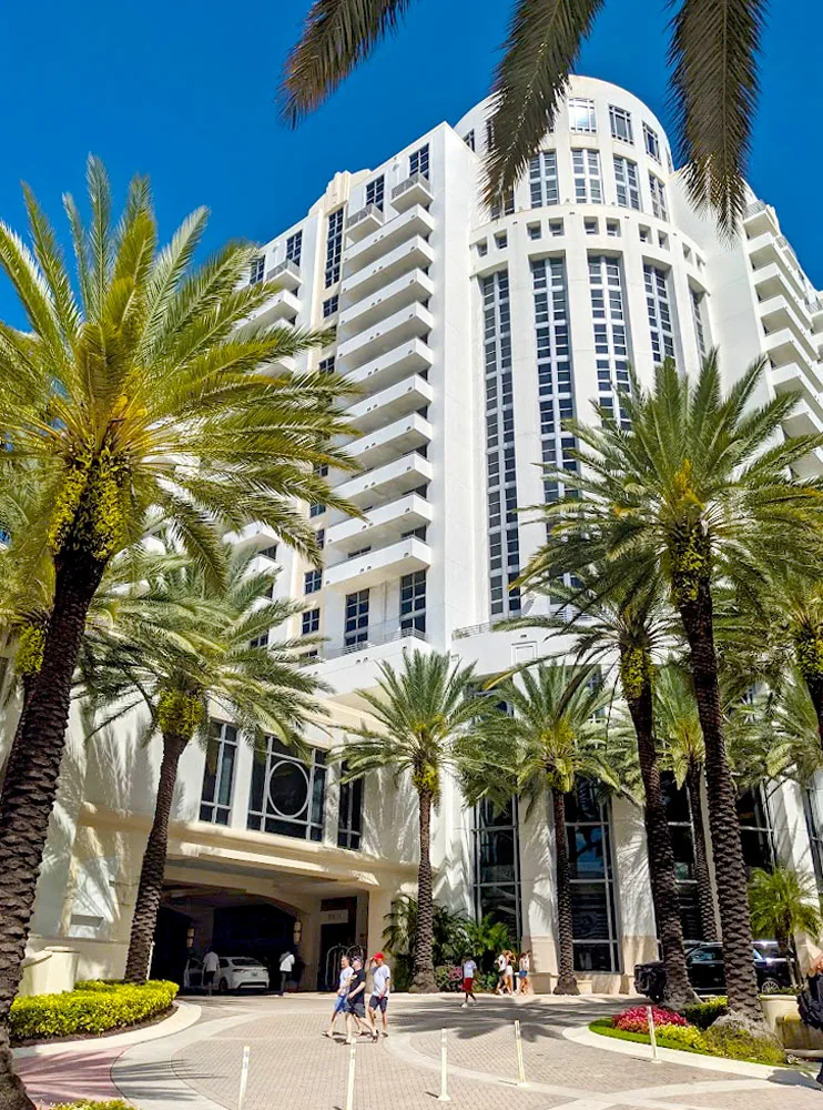 a stark white high rise building with art deco style entryway for cars to drive under. People are walking in front of it, looking like ants in comparison to the size of the hotel. Even the palm trees only make it halfway up the giant hotel. 