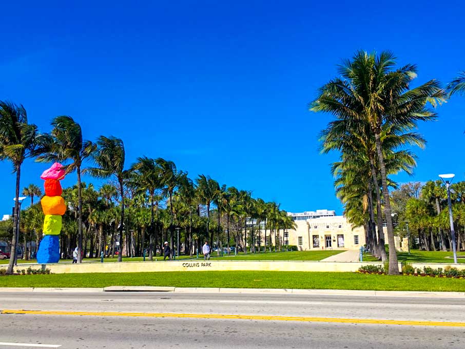 the road with collins park  across the street. There's a standing rock sculpture, each rock is standing on the other but each is painted a different vibrant neon colour, The bottom is blue, neon yellow, orange, red and pink. There are palm trees lining the way to the museum building at the end of the park. 