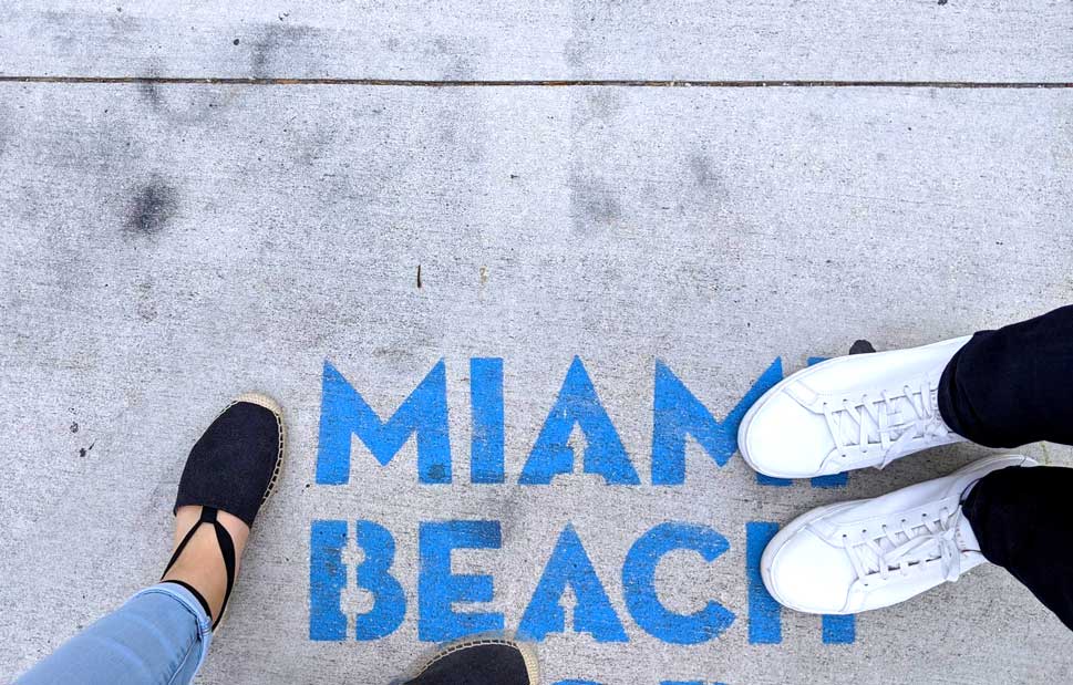 two pairs of legs and shoes photographed from above on the sidewalk in miami. Spray painted in blue  in a stencil font it says Miami Beach. 

On the left are a womans feet wearing black espadrilles and light blue skinny jeans. On the right, covering the text are perfectly white sneakers and black pants. 