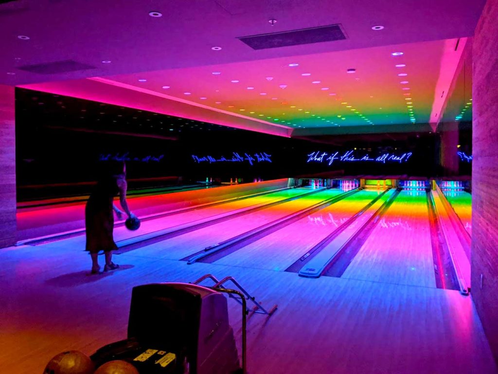 a neon light bowling alley with a rainbow of colours. There are four alleys with pins at the end. One woman's silhouette is seen with a bowling ball in her hand about to lunch it down the lane.