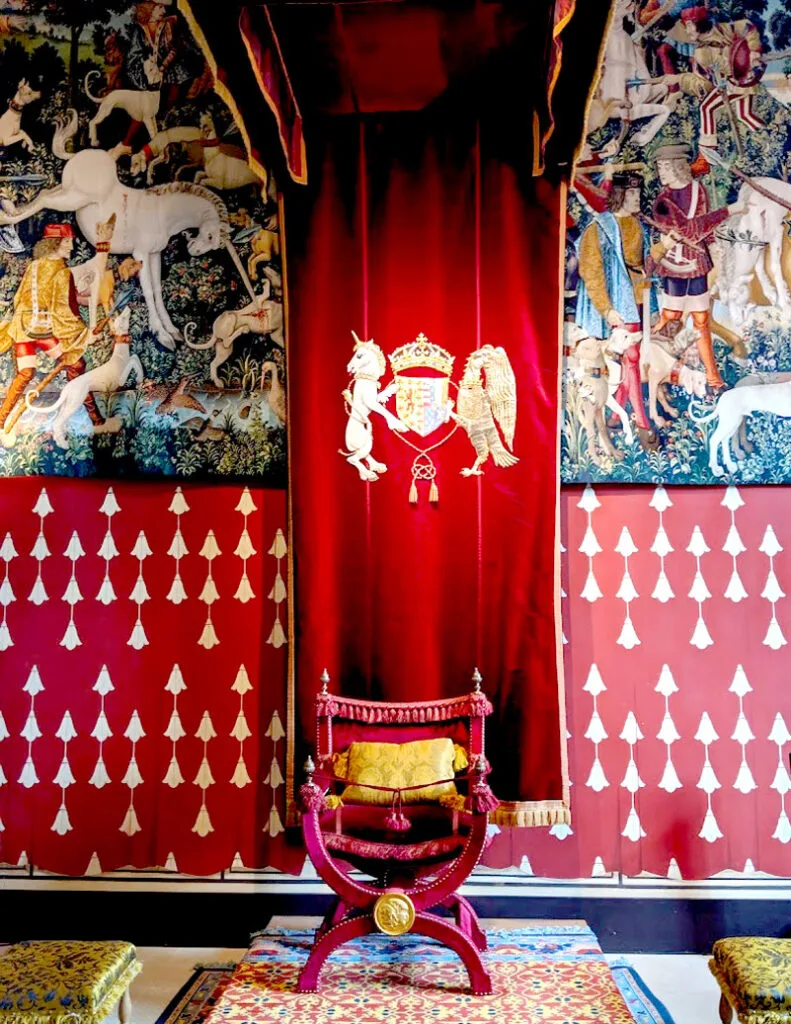 an example of the restored decor, tapestries, and art in stirling castle in Scotland. The floor is finished with a carmet in the middle (it has a red, yellow, and blue pattern). On the carpet sits one very unique chair - unlike any chair from the modern world. The legs create almost the coco chanel logo. An arched C facing downward creates the legs. Connecting to the legs is another C facing upwards. This hold the armrests. The chair is a deep burgundy royal red, with golden yellow accents, and a fluffed pillow in the middle of the chair. 
Behind the chair is a wall. The wall is separated a top and bottom. The bottom is a red wallpaper with white tassles as the print. The top is a painted tapestry  of a typical medieval times scene with horses, men, and dogs. In front of the wall hangs a red velvet tapestry with the Scotland emblem in the middle (a lion and a unicorn holding the Scottish crest). 