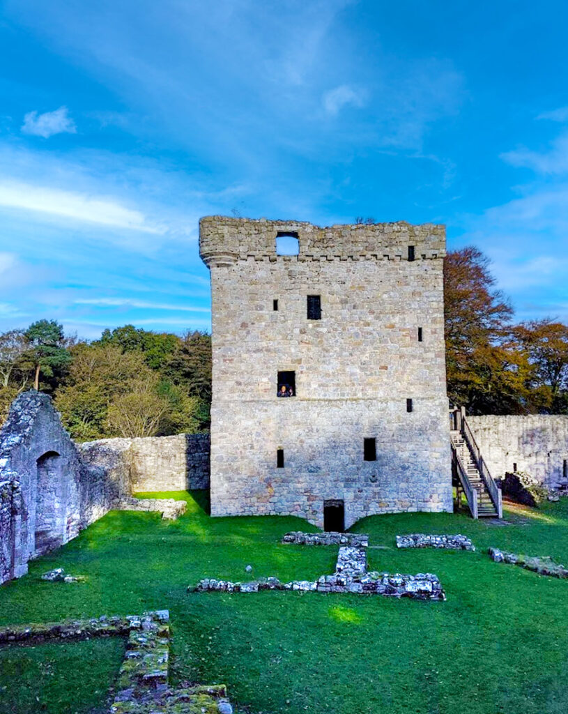 a scottish castle scene: some stone piles on the green grass courtyard. Surrounding this courtyard is a stone wall, with a triangle shape doorway out. At the end, right in front of you is a 5 story tower. Inside on of the windows you can barely make out two people standing. Wooden stairs lead up to the third story. The windows are randomly placed and are all different sizes. There is one a door in the middle on the bottom floor.