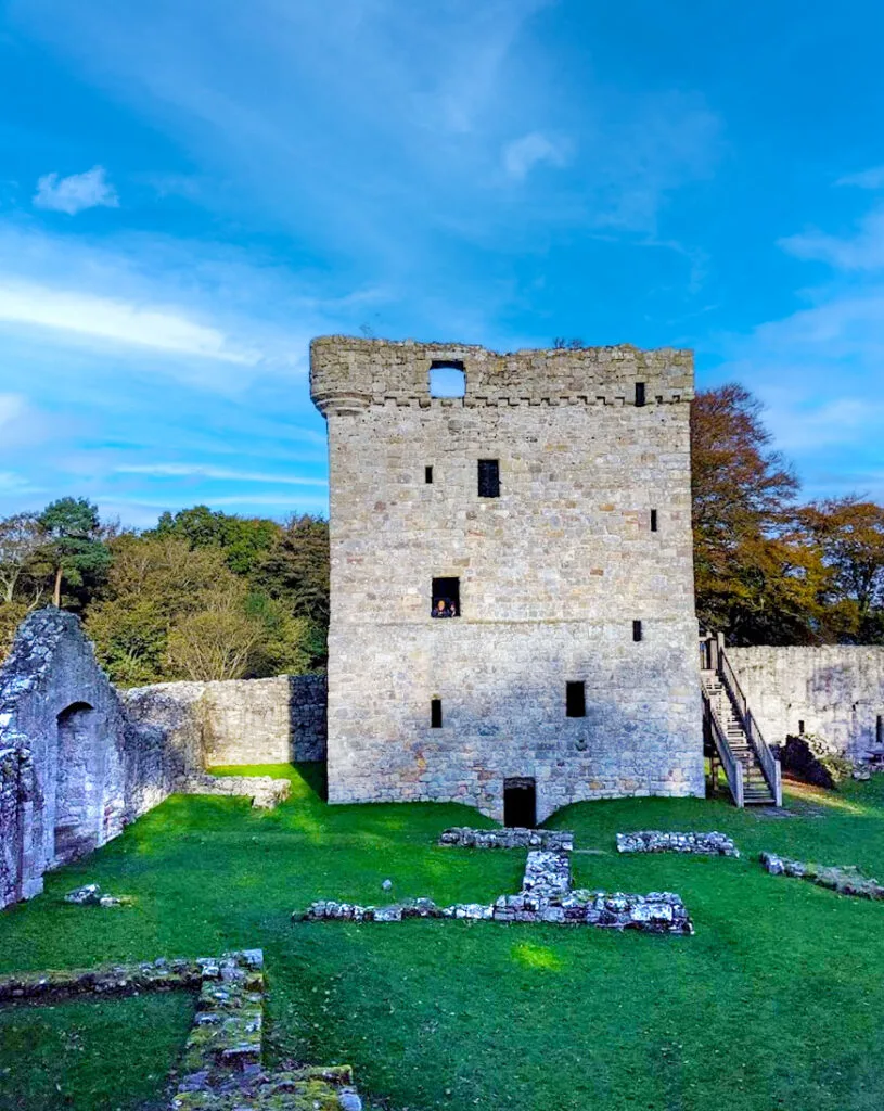 a scottish castle scene: some stone piles on the green grass courtyard. Surrounding this courtyard is a stone wall, with a triangle shape doorway out. At the end, right in front of you is a 5 story tower. Inside on of the windows you can barely make out two people standing. Wooden stairs lead up to the third story. The windows are randomly placed and are all different sizes. There is one a door in the middle on the bottom floor.