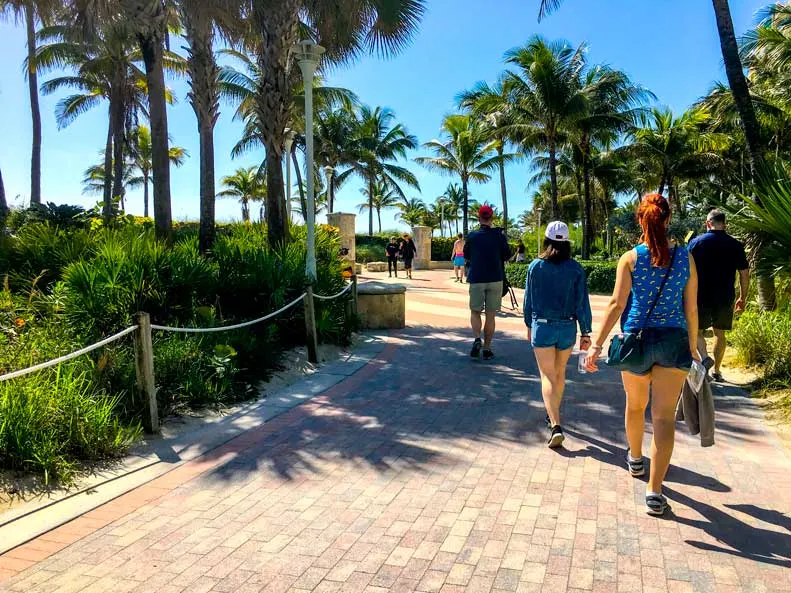 a family with their backs to the camera walk along the stone path boardwalk. There are bushes and palm trees on either side of them, shielding them from both the road and the beach. 