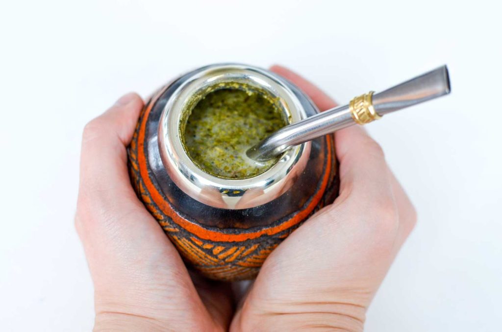 two hands cup a traditional yerba mate gourd between them. It looks like the hands are holding on tight to consume all the gourds warmth that eminates from the hot yerba mate tea inside. The tea inside looks like thick green chunky liquid. Sticking out is a stainless steel straw.