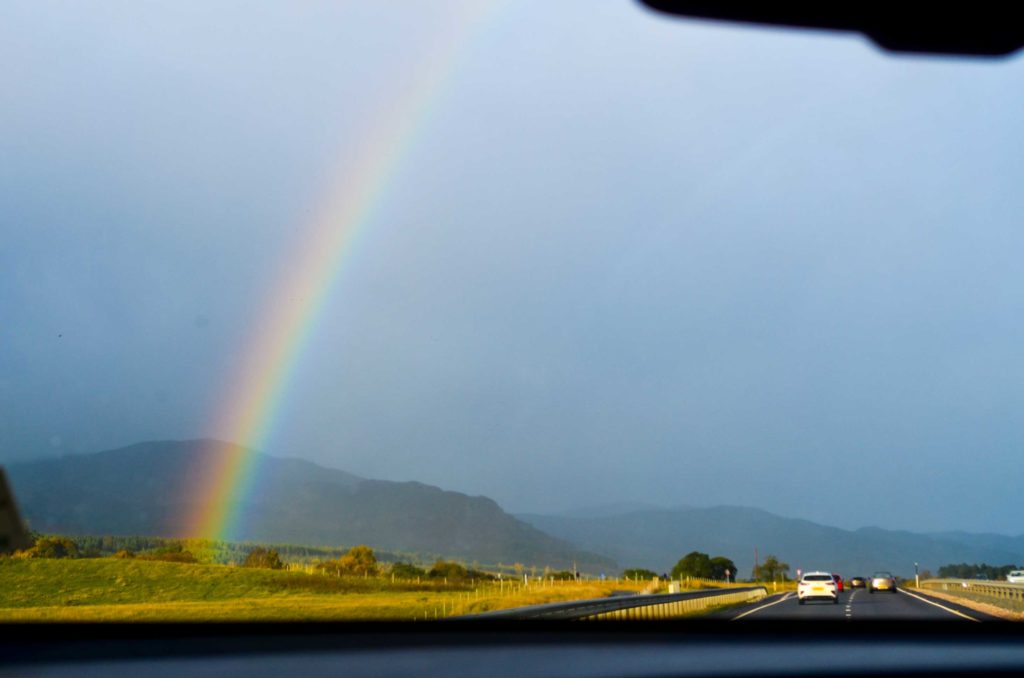 driving in scotland, the views are magnificent. Out the front windsheilf is the road ahead, looking rather ominous in the distance. The sky is hazy, adding a layer in front of the mountains in the distance. But head of them is a perfect rainbow coming from the ground and meeting the sky