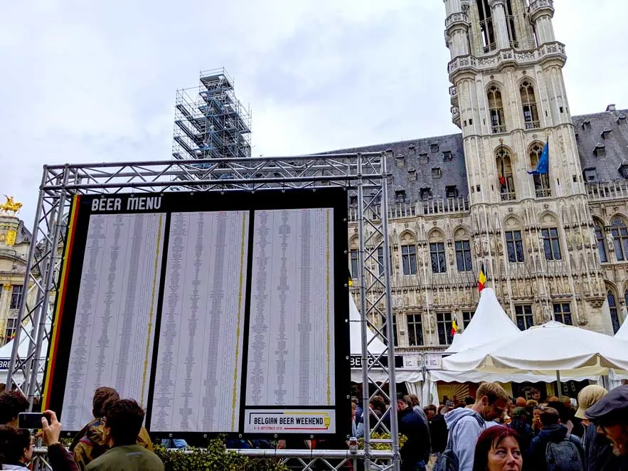 belgian beer weekend brussels festival taking place in grand place (you can see a bit of the city hall gothic building in the background. But covering it is a giant billboard with a list of all the beer vendors at the festival. People are walking in front of it, and behind it, below white tents that have been set up in grand place for the festival.