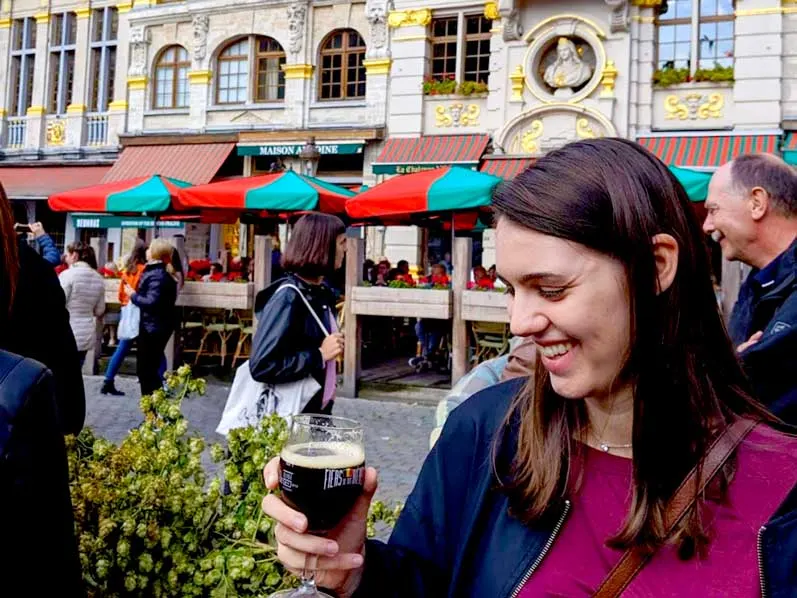 a woman smiling at a beer glass with a dark brown beer inside. 

There are green and red umbrellas in the background in the grand place in Brussels. 