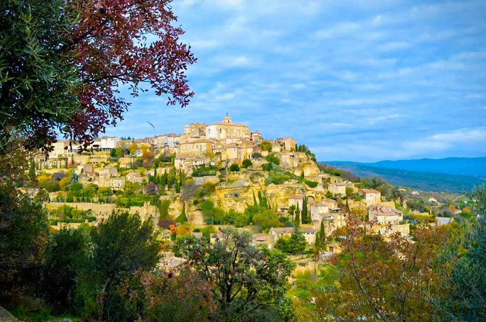 The jaw dropping view of the town of gordes in the south of france from a distance. You can see the entire medieval town built on top of a the hill leading all the way down to the valley. Trees are intertwined with yellow stone buildings. 
A tree blocks the left side of the frame, but you can tell it's autumn because the tree is in the middle of changing from green to a deep red.