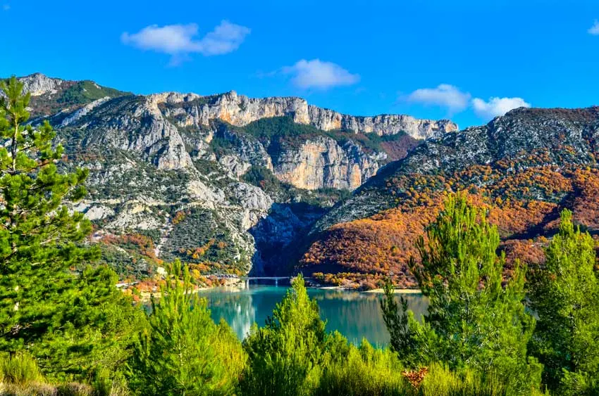 A peaceful photo of the gorge de verdun in the fall. Evergreen trees line the bottom of the frame. Peaking through beyond the trees, you see a crystal blue lake, with the reflection of the tall rocky mountains above. The bottom of the mountains have some trees, some are still green while others have burnt orange leaves already. 