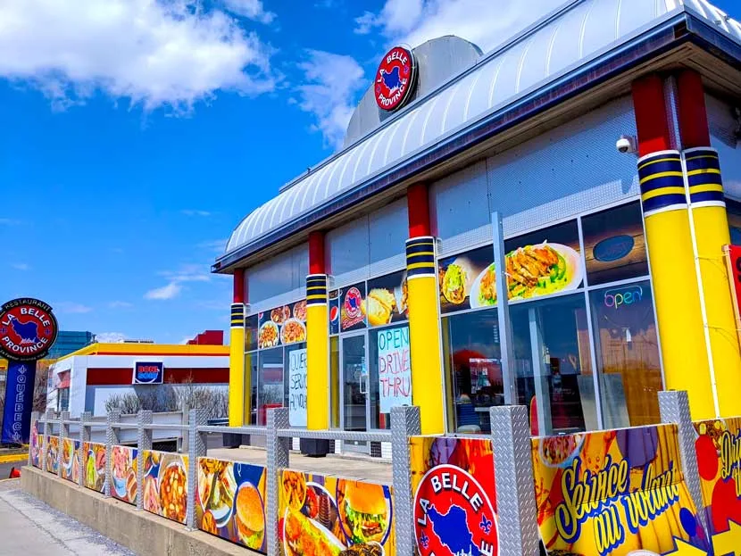 outside of belle province, it looks like a building from the 50s outer space renders. The roof is silver aluminum, the siding is mostly windows, except for bright yellow columns, with some navy stripes in the middle, and red at the top. There's a gate surrounding the outside patio, all with photographs of the food on the menu. In the distance is La Belle Provence red and blue highway sign.