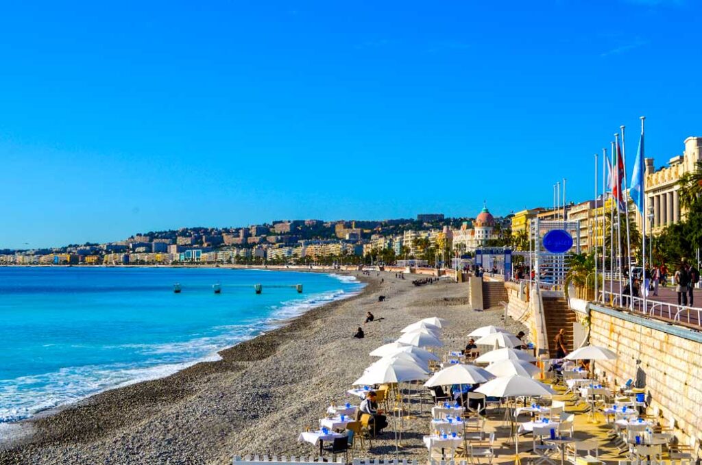 a peaceful photo of the quiet nice beach in the fall in the south of France. The water is slowly coming in to the beach. There's enough space before it reaches umbrellas and chairs set up. Behind the chairs is a concrete wall separating the street. above. You can't see the street, but you can see the shops and apartments all along the way.