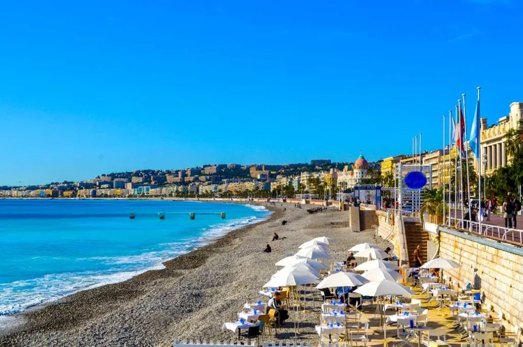 a peaceful photo of the quiet nice beach in the fall in the south of France. The water is slowly coming in to the beach. There's enough space before it reaches umbrellas and chairs set up. Behind the chairs is a concrete wall separating the street. above. You can't see the street, but you can see the shops and apartments all along the way.