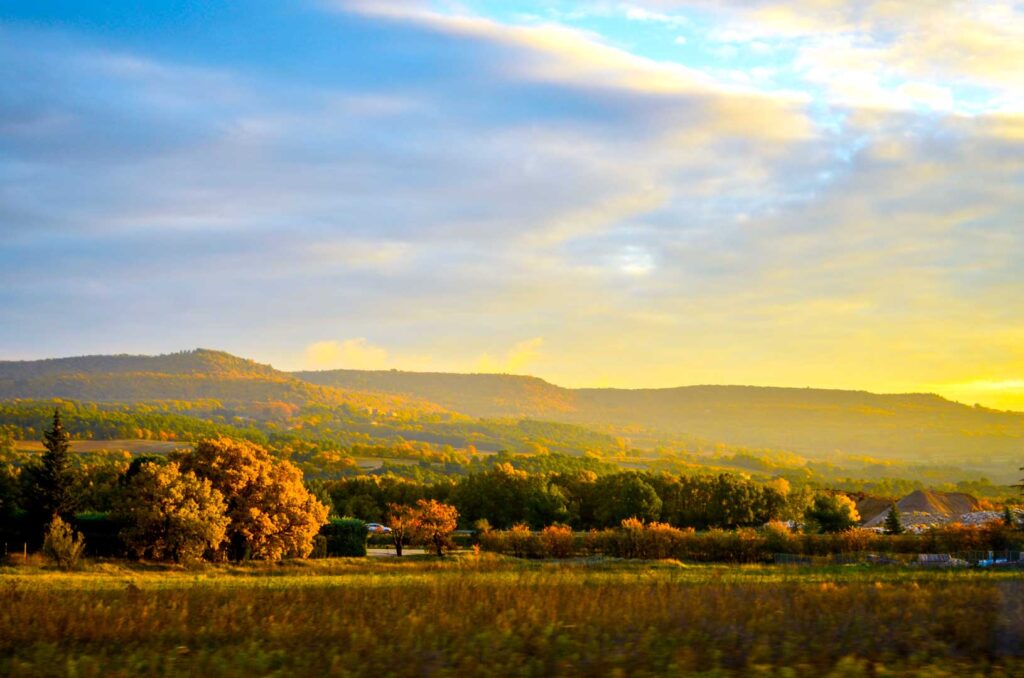 A romantic Provence sunset in the fall. The sky is a bit more than half the photo, the clouds are whispy across the sky, but the gradient of colour changes from light blue in the right corner, to a darker blue in the left. As you follow the sky down towards the middle, a golden yellow is added, before the yellow completely takes over. The middle of the photo are the mountains, covered in fall trees and the tree lead all the way to the front of the photo before a Provence field takes over the bottom of the photo. The trees are a mix of a dark green, yellow, and burnt orange.  