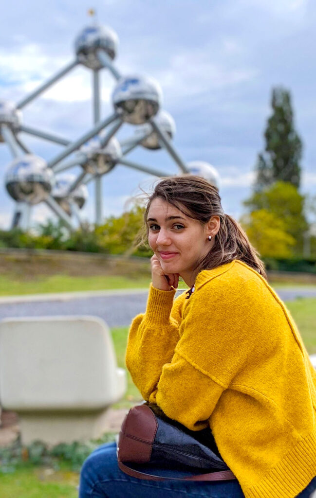 a woman wearing a bright mustard yellow wool knit cardigan, she's sitting on a bench, with her head resting on her hand. She's smiling, but without showing any teeth. The background is slightly blurred, but you can easily make out the large metalic structure in the background: the Atomium, with the belgian flag on top. 