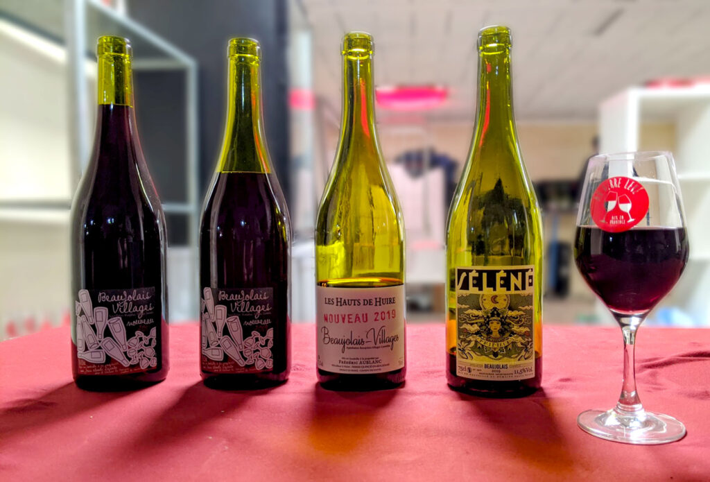 four wine bottles and a wine glass sit on a table with a red table cloth. The background is blurred. Each wine bottle has a different amount of wine still inside, they are green bottles holding red wine. From the left, the first two wine bottles are the most full and are the same wine: Beaujolais Village. Next  is a bottle half finished with a white label: Les Hauts de Huire Nouveau 2019, Beaujolais Village. And finally, a bottle with a only a sip left, is Selene Beaujolais.