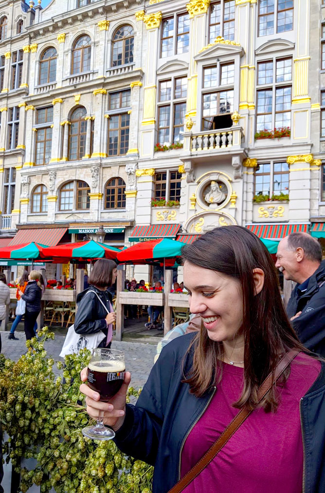 a woman smiling at a beer glass with a dark brown beer inside. There are green and red umbrellas in the background in the grand place in Brussels.