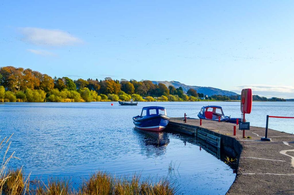 two boats are attached to a permanent paved dock in the lake. One boat faces you, while the other one on the opposite side of the dock, faces outward towards a completely tree covered island in the middle of the lake.