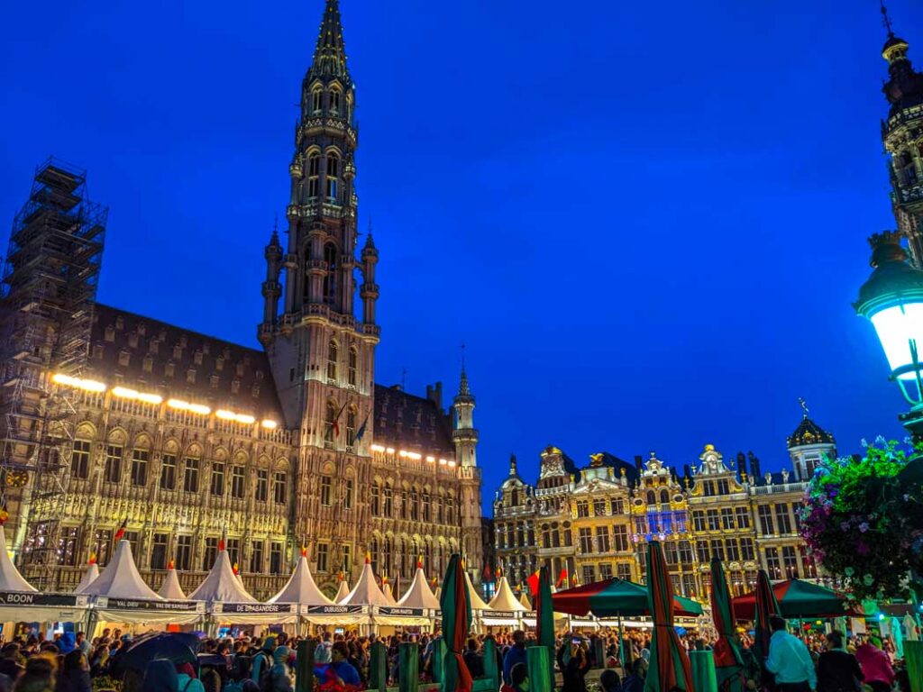 brussels grand place at night during the beer festival in september. The sky is completely clear, and it's a royal blue. The tower of the buildind goes high into the sky. It makes the golden decorated buildings next to it seem small. The middle of the square are filled with white tents and thousands of people that came to the festival. 