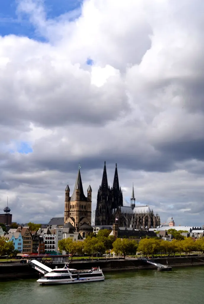 The view of cologne city across the rhine river. The river is a dark teal colour, and there's a few cruise boats docked on the side. Trees line the path, and high above the trees and every other building are two dark towers - the cologne cathedral. The clouds are huge and scary, turning more grey the closer they get to land.