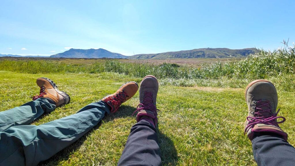 a wide lens photo of peoples legs and feet sticking up as they sit on the grass in Iceland. There's a mountain in the distance (likely a volcano). It's the legs of a man and a woman, although they are wearing the same shoes: columbia newtons. The mens are a classic like tan colour with red accents. The womans are olive green with a deep pink accent,