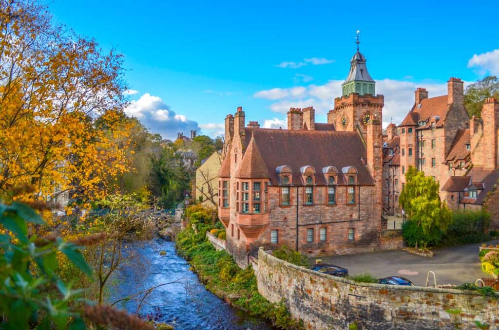 deans village in Edinburgh in the fall. A romantic photo of a red stone building with turrets and bay windows over looking a rushing river below. Some trees overtake the left side of the frame, with yellow leaves. 