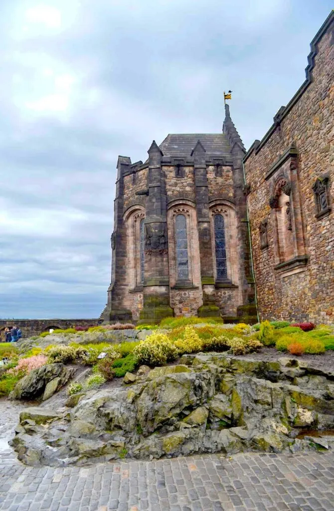 a striking vertical photo of a tower in edinburgh castle. The natural rock ground is covered in moss, it leads right into the stone building of the castle. It's darker grey the ground, it's either raining or just rained. The sky is also not inviting, completely covered by thick clouds