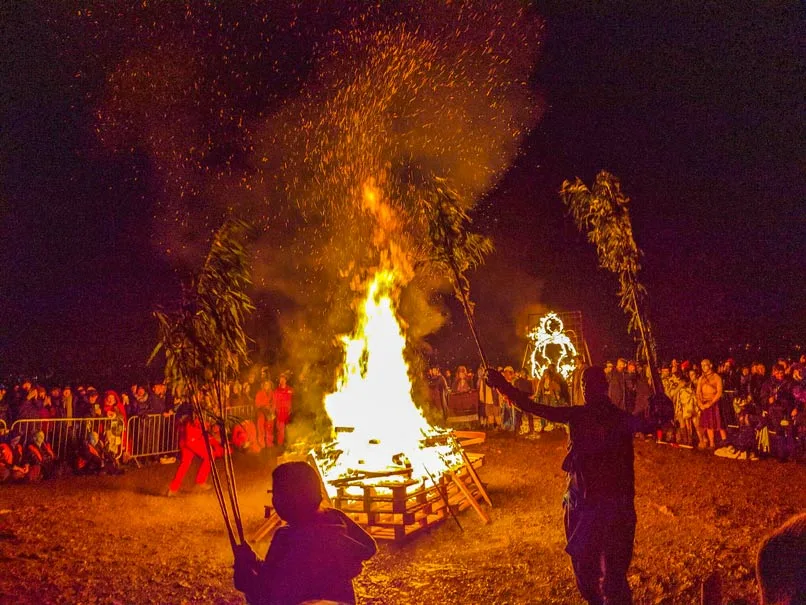 the incredible edinburgh beltane samhuin  fire festival on halloween in october. This is the grand finale of a giant bonfire in the centre and people watching from a safe distance. Participants in the festival are holding large branches with leaves around the fire. It's pitch black in the night (it was also raining, but you can't see that in the photo). The only light comes from the giant fire in the centre illuminating the scene to be orange and adding shadows to others. 