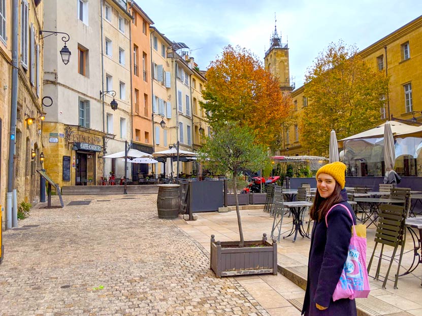 a woman stands on the right side of the photo looking back at the camera. She's wearing a mustard yellow knit hat, a wool jacket, and carrying a bright pink tote bag over her shoulder. She's standing in the middl eof a quiet square in aix en provence in the fall. The bulidings are all attached, wrapping around teh empty square. Each is painted a different shade of yellow. You can't see where the opens, because a green and a red tree are at the end of the courtyard. Poking above the trees looks like the steeple of a church. 