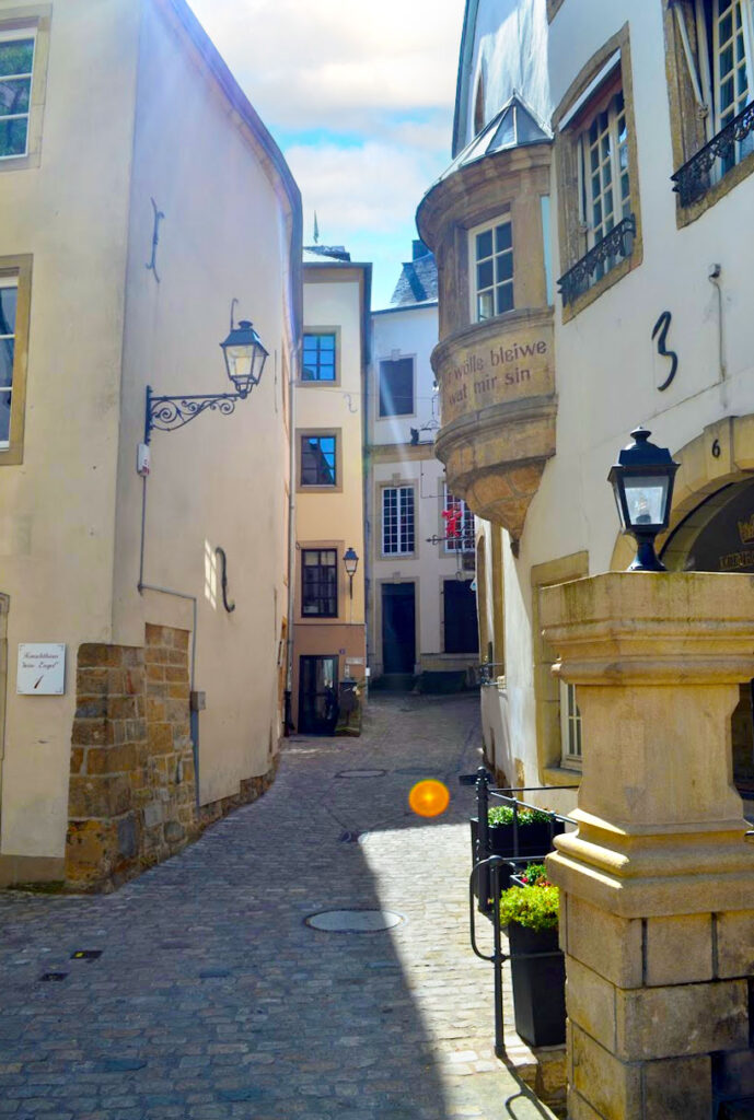 an empty street in Luxembourg luxembourg in the fall. The light is creeping through the taller buildings and casting shadows on other ones. The architecture looks more medieval, none are brick but flat facades in light orange and white. There is one bay window sticking out from the second floor on the building in the foreground