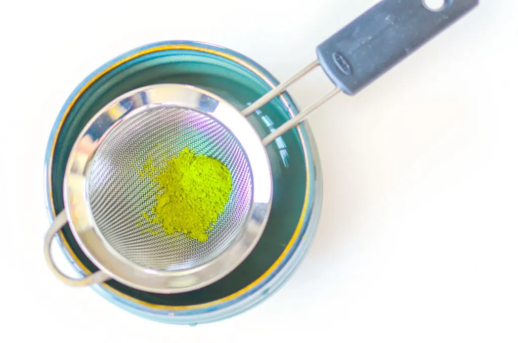view from above. a white table so you can focus on the teal matcha bowl with a sift leaning on the rims and bright green matcha powder inside waiting to be sifted