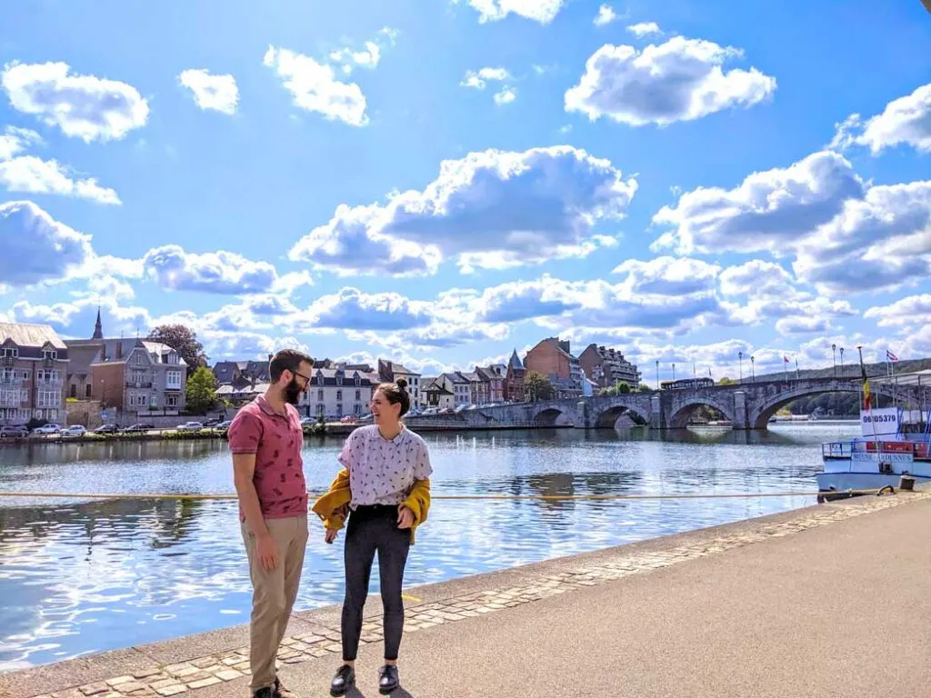 overheating in namur september heat. Along the river meuse, a couple stands on the sidewalk, they're looking at each other, smiling. The womans hair is up in a tight bun, she has a thick mustard yellow knit cardigan, but it's off her shoulders as it's hotter than she expected. The are both wearing a t shirt and pants. Behind them, on the other side of the river are some homes and shops, you can see a bridge crossing the river connected both sides. 