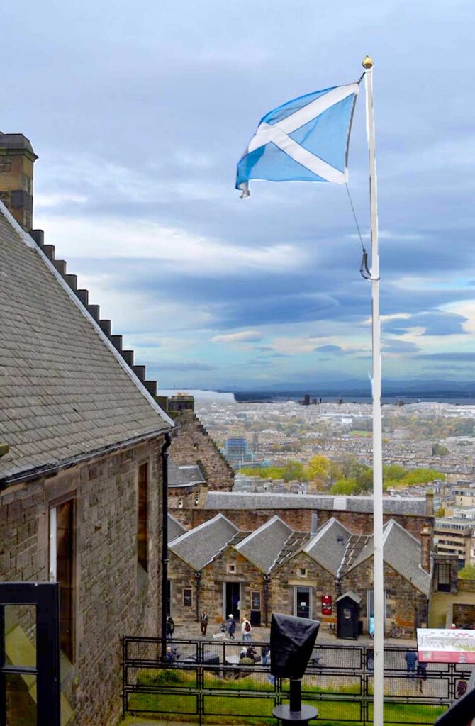 a scottish flag blowing in the window above edinburgh from edinburgh castle. There's an old timey stone hime next to it on the same level. But then you dip to a lower level, new town edinburgh you can see the front facade of the homes on princes street. The sky is cloudy and ominous above