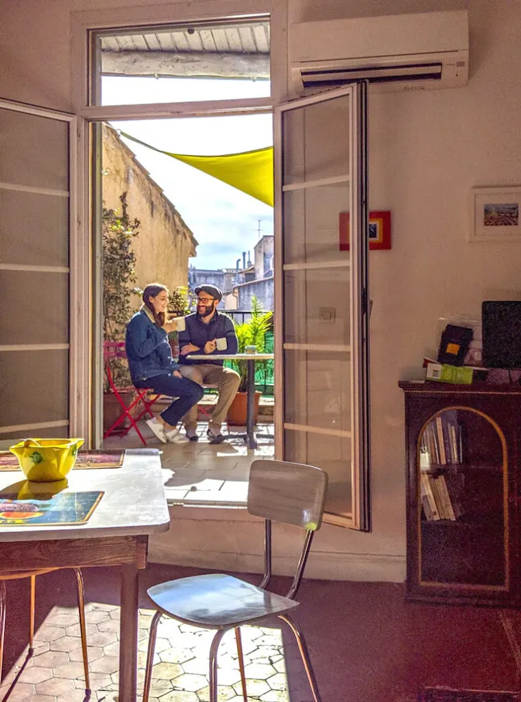 a snapshop into a couples life in the south of france. The photo is take inside an apartment, you can see a small kitchen table and a pulled out chair. The floors are red tiles. Behind, are two open patio doors. It's letting in some sunlight into the apartment. Outside is a couple, wearing jackets, and a har, sipping on some warm tea trying to warm up in the fall sun in the south of france in aix en provence