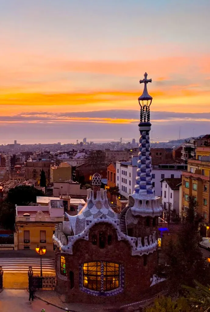 a beautiful scene of sunrise over park guell. The sky is a mix of sun and clouds, creating a mix of blues oranges, pinks, and yellows across. You see the skyline of barcelona city in the distance. In the front are the gates of park guell that look like a fun candy shop with a cross on a spire  on top of a small cottage. 