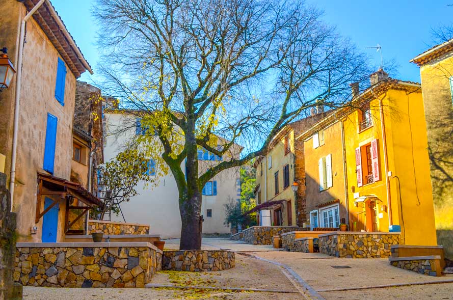 a very quiet town in the gorges de Verdun in the south of France in the fall. The homes are attached together, painted in golden yellow, pink, and orange colours, they look even more golden because the light is hitting the homes on the right, while the others are in complete shade. Raised stone entryways are in front of all the homes. There's only one tree in the courtyard, and the leaves have mostly fallen off, leaving the branches bare.