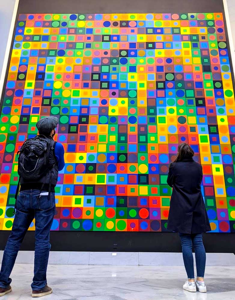 two people stand with their back to the camera, admiring a larger than life floor to ceiling piece of art by vassarely at the art museum in aix in the fall. The art is a large rectangle filled with different coloured squares and circles, it doesn't seem as though there's a pattern to the colour and shapes. 

The two people are wearing jackets and hats with long pants.