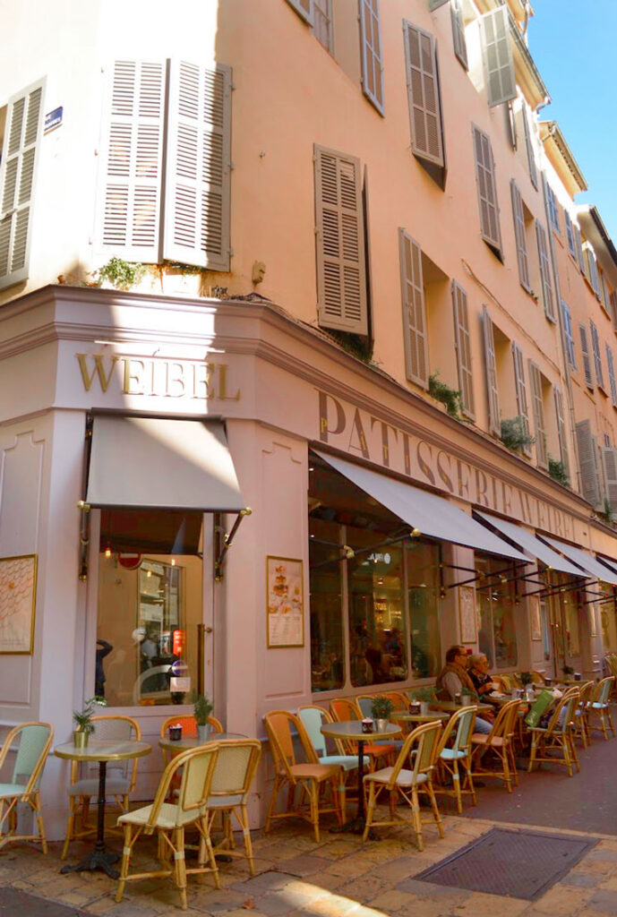 the outside of weibel pattiserie in Aix en provence. It's a romantic facade, with a table and two chairs lining the outside wall. There are lavender awnings above the windows and flowers on the window sill. The name is in gold letters above the awning. There are two more stories above the awning, must be apartments. The building is a light pink stucco. There's only one table with people sitting outside