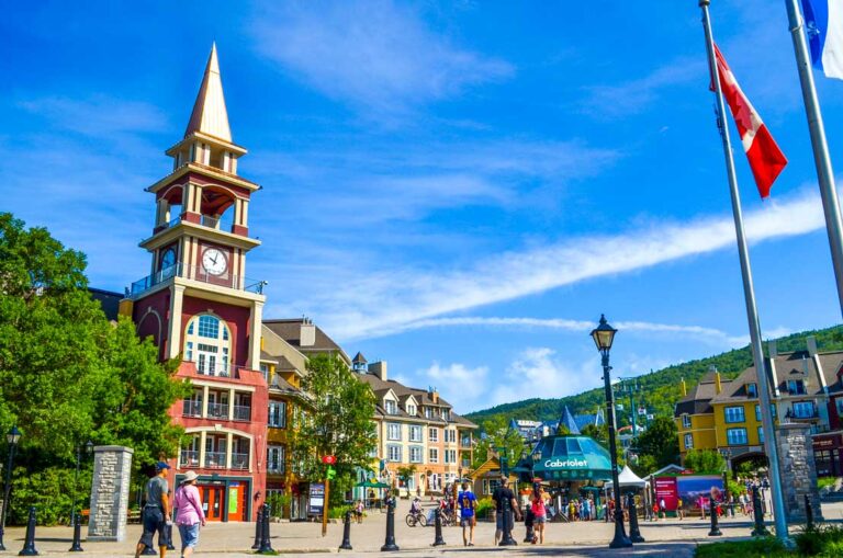 Mont-tremblant-in-the-summer-day-trip-from-montreal a clock tower at the front with hotel buildings behind you can see the entrance to the cabriolet and people walking around