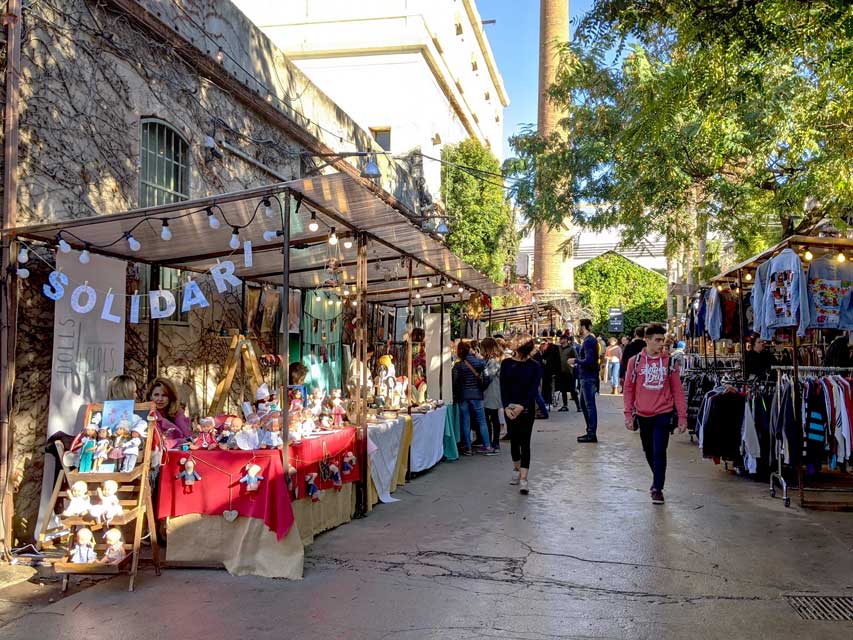 barcelona holiday market, pop up shops are set up on either side, creating a corridor for people to walk through ad peruse. There's a shop with sweaters, another with trinkets. Trees are looming above, creating shade.