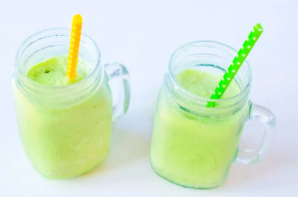 two frozen matcha slushies on a white table in mason jars with handles. They're bright green thick drink. There's a yellow and green polka dot straw in each one.