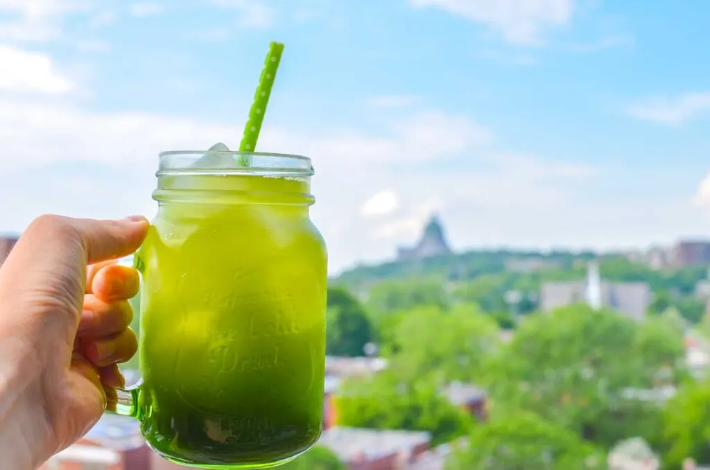 matcha honey iced tea summer recipe. A hand holding up a green iced drink in a mason jar with a green polka dot straw sticking out. Blurred in the background are bright green trees and buildings on a nice summer day