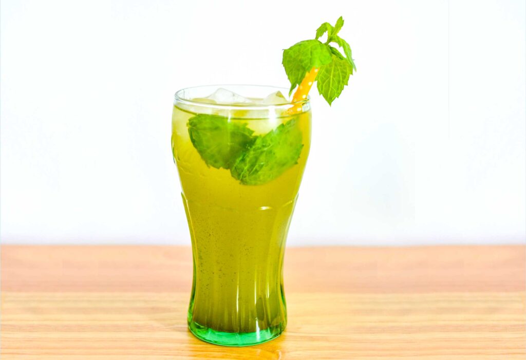 matcha mint lemonade  on a wood table with a white background. It's a olive green drink with floating fresh mint leaves. There's also mint leaves sticking out of the yellow polka dot straw in the iced drink.
