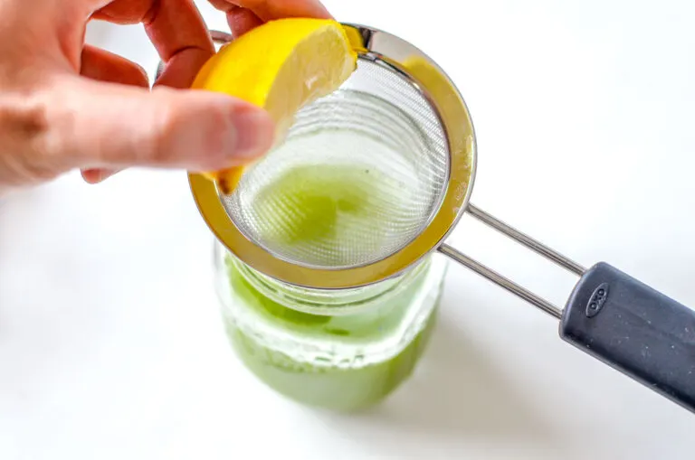 a person is squeezing a lemon slice over a mini strainer placed on the lid of a mason jar already filled with green drink (matcha green tea).