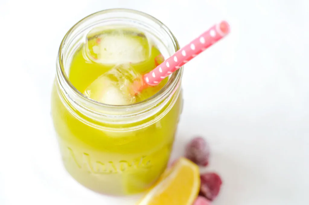 matcha raspberry lemonade from above. A greenish yellow drink filled with ice cubes in a mason jar. There's a pink polka dot straw sticking out. Next to the jar are some blurred raspberries and a lemon wedge