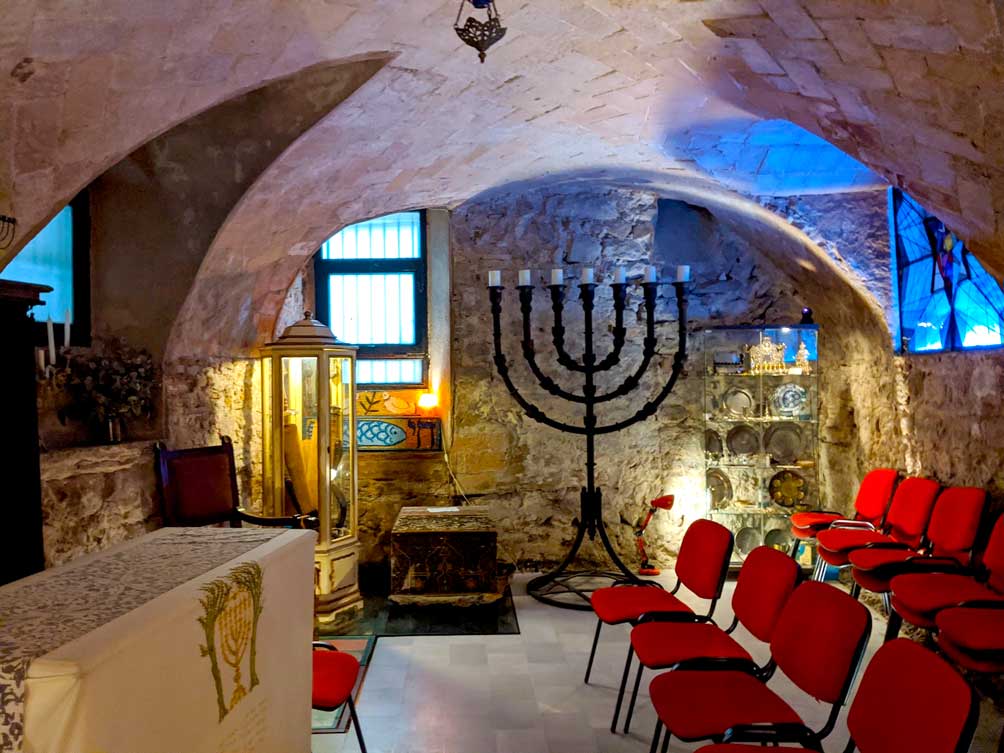 the old jewish synagogue in barcelona is underground. There's a standing old brass menorah in against the stone wall in the back. The ceilings are so short, they are curved above. There's a glimpse of where windows are, just from a blue glow of light from either side of the room. There are two rows of red chairs on the right and a stand on the left side