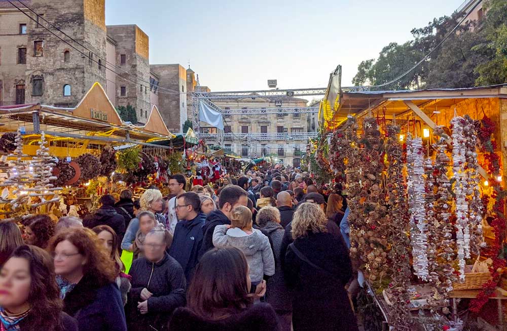 a completely packed market in barcelona spain in the gothic quarter on constitution day weekend. There's barely any space between the people walking in every direction and the booths set up on the sides with christmas lights to sell their merchandise. The old buildings of the gothic quarter rise above the booths and people and you can see them in the distance.