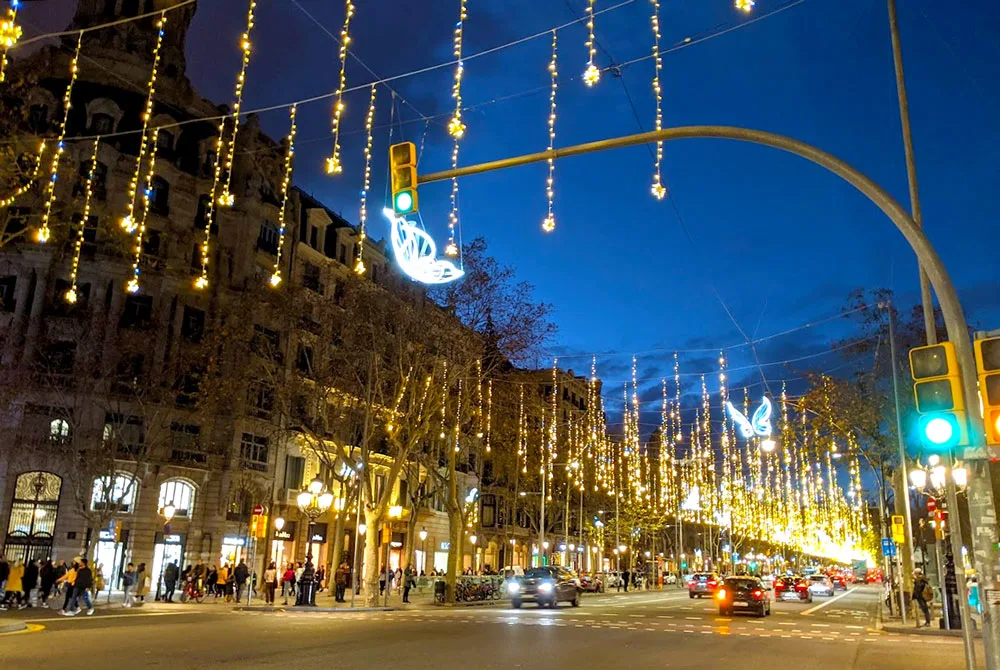 the view down passieg de gracias at night. The sky is a beautiful darker to lighter royal blue gradient. The colour getting lighter as it gets closer to the city lights. The street is beautifully lit up with hanging yellow fairy lights and butterfly lights across the middle. It looks like they're floating in mid hair. Cars drive down the street respecting the green light.