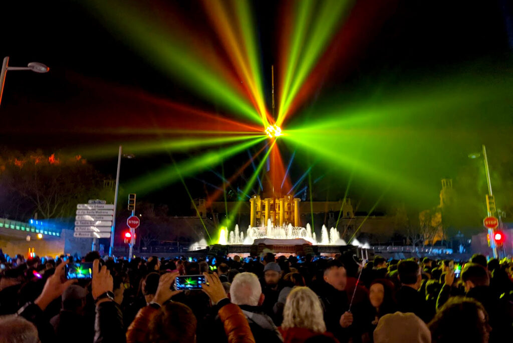 Plaça d'Espanya in barcelona on new years eve. Standing behind a crowd of people, some are trying to leave, but most are facing the spectacle in front of them, and you see them holding him their phones to take pictures. At the far end, among a dark background, is a fountain and a light show coming from a ball that seems to be floating in the air. Rays of green, yellow, and red light shine across almost reaching the edge of the photo. 